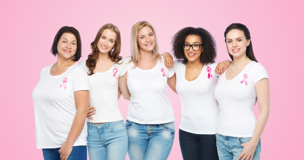 5 women stand in front of a pink background. They wear white shirts and have a small pink ribbon pinned to their shirt. Tags: Breast Cancer Awareness, 3D Mammo, Mammography, Call 844.414.3627 to book your appointment today.
